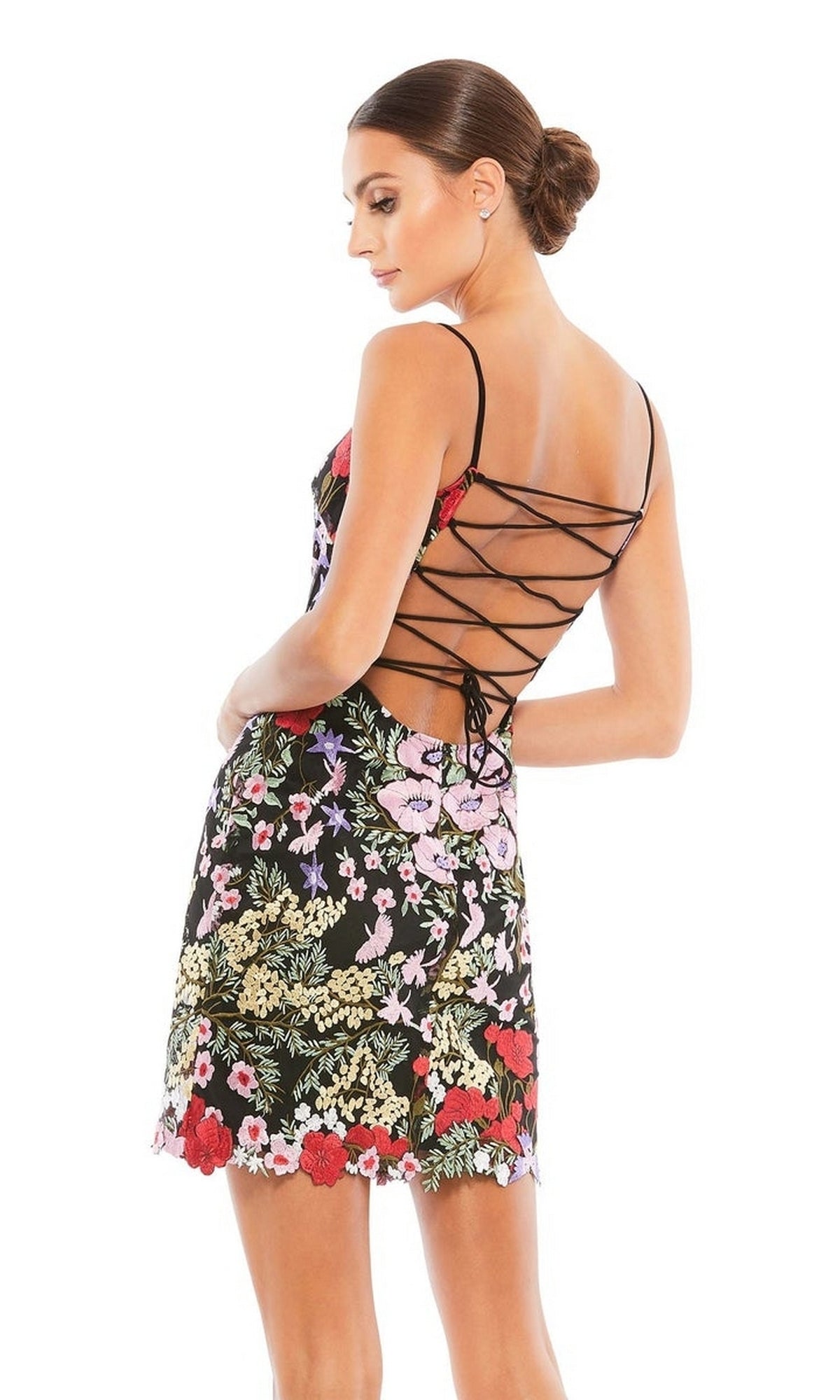 A woman in the Mac Duggal 67459 short floral-print party dress with spaghetti straps stands sideways, her hair styled in a low bun.